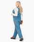 A woman modeling a slate blue sleeveless jumpsuit over a white long-sleeve top, wearing brown platform shoes and carrying a small brown shoulder bag, set against a plain white studio background.