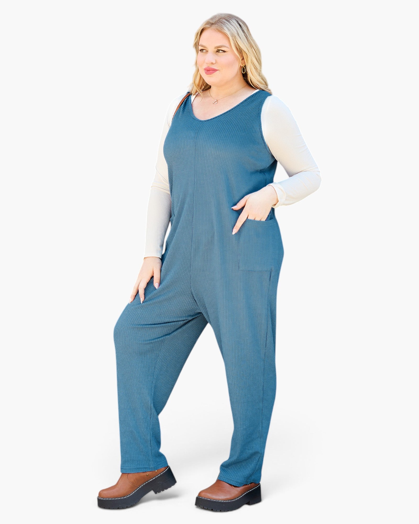 A woman modeling a sleeveless slate blue jumpsuit worn over a white long-sleeve top and brown platform shoes, photographed against a clean white studio background.