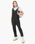 A woman modeling a pair of black sleeveless overall-style pants with a white t-shirt underneath, carrying a beige shoulder bag and wearing white platform lace-up shoes against a light gray studio background.
