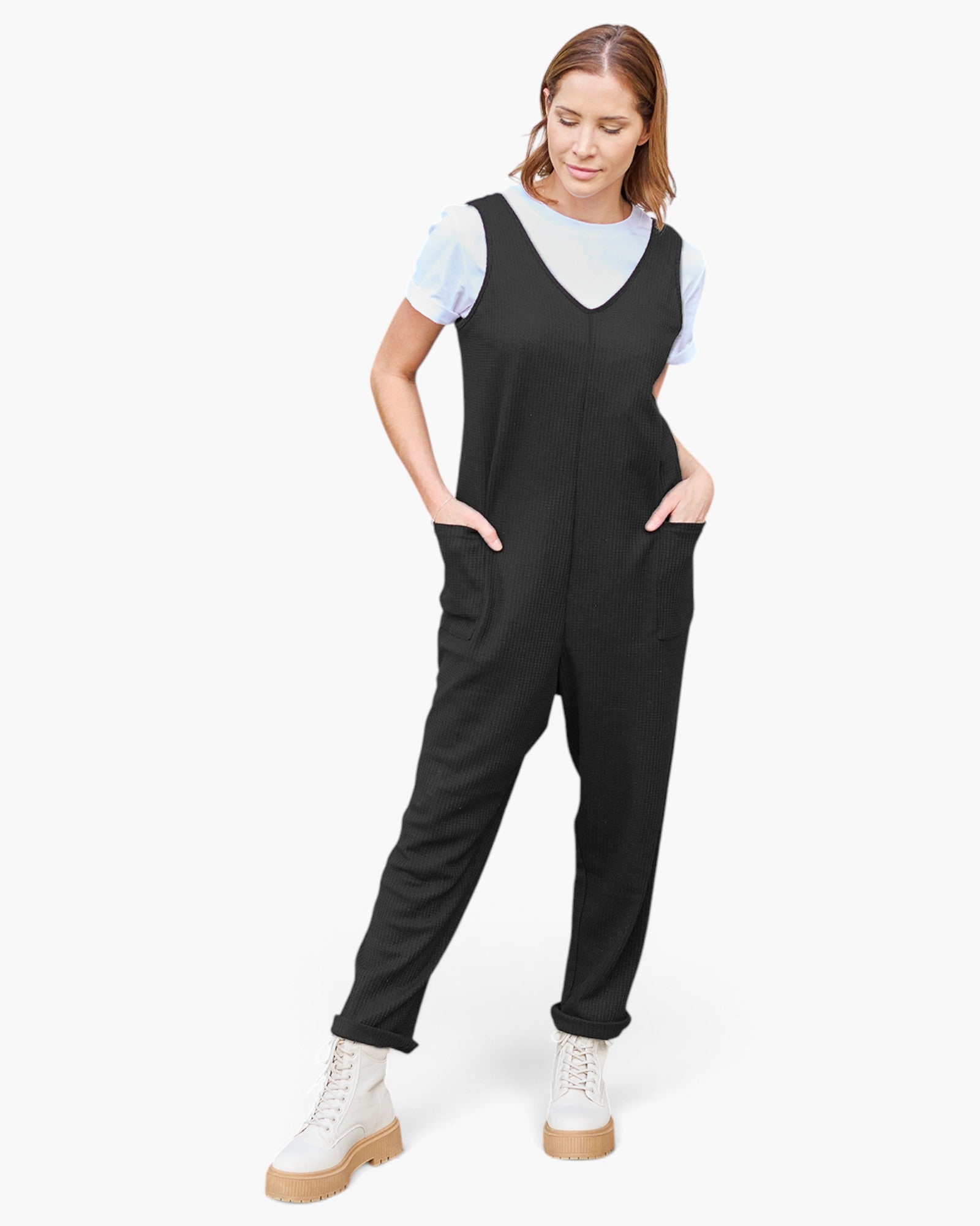A woman modeling a black sleeveless jumpsuit with a V-neck and side pockets, worn over a light blue short-sleeve t-shirt and paired with white lace-up boots, on a light gray background.