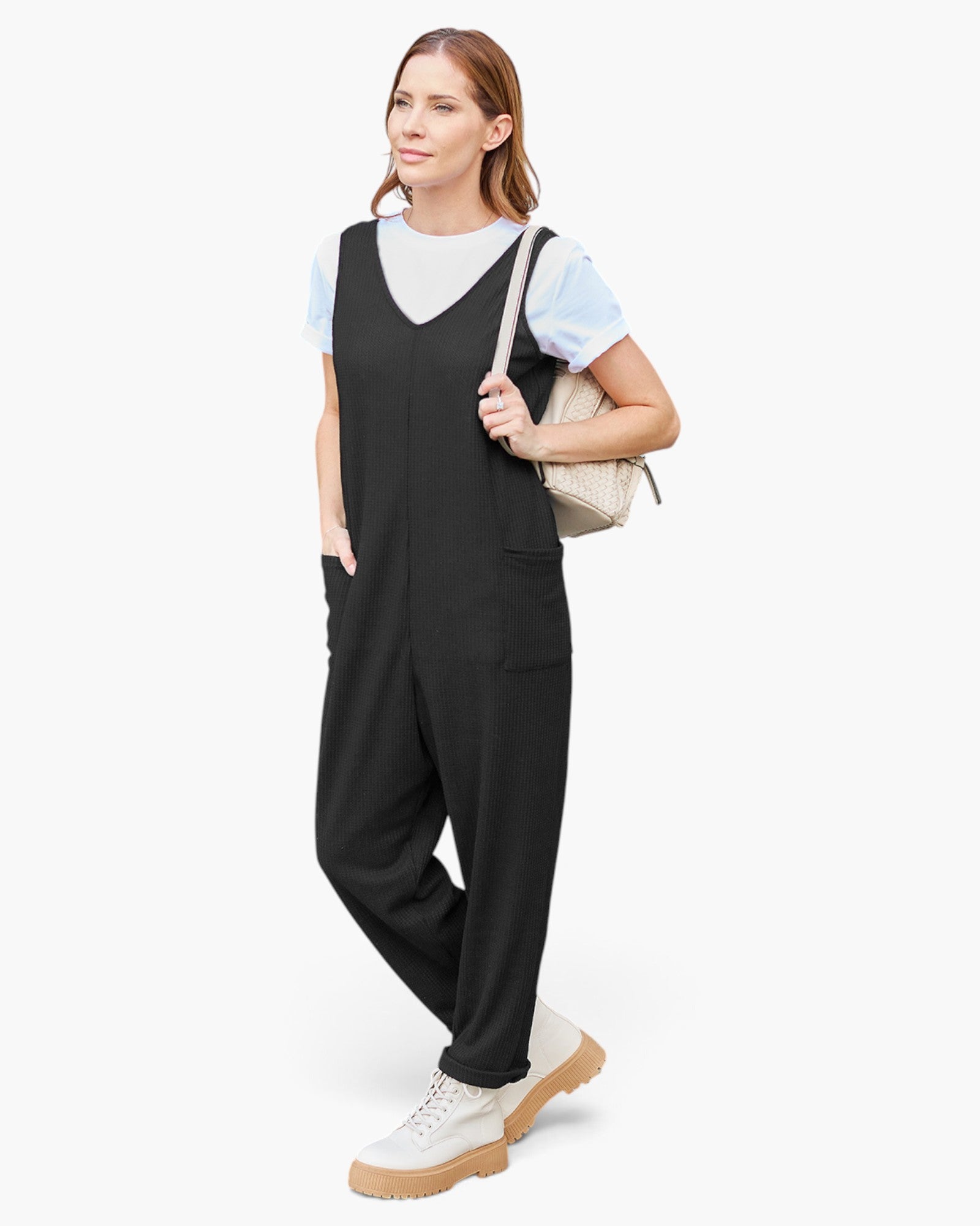 A woman modeling black wide-leg overall jumpsuit with a white crewneck t-shirt, white sneakers, and a beige shoulder bag against a plain white background.