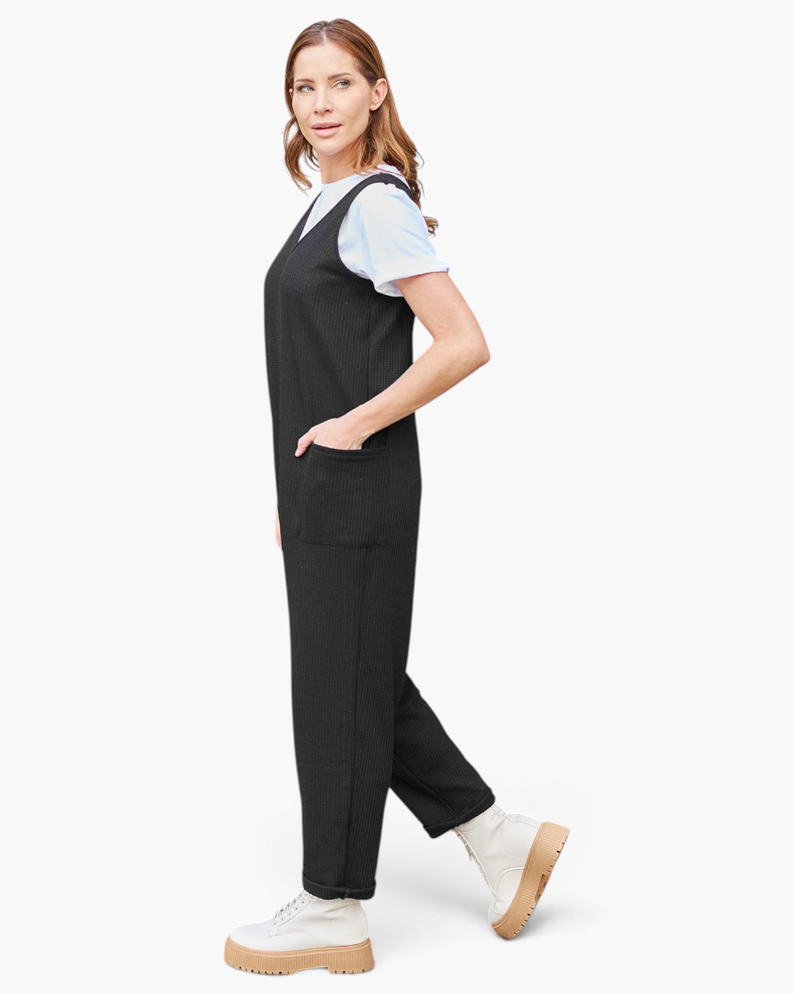 A woman modeling a pair of black scrub-style overalls with a white short-sleeve top underneath and white platform sneakers, photographed against a clean light gray studio background.