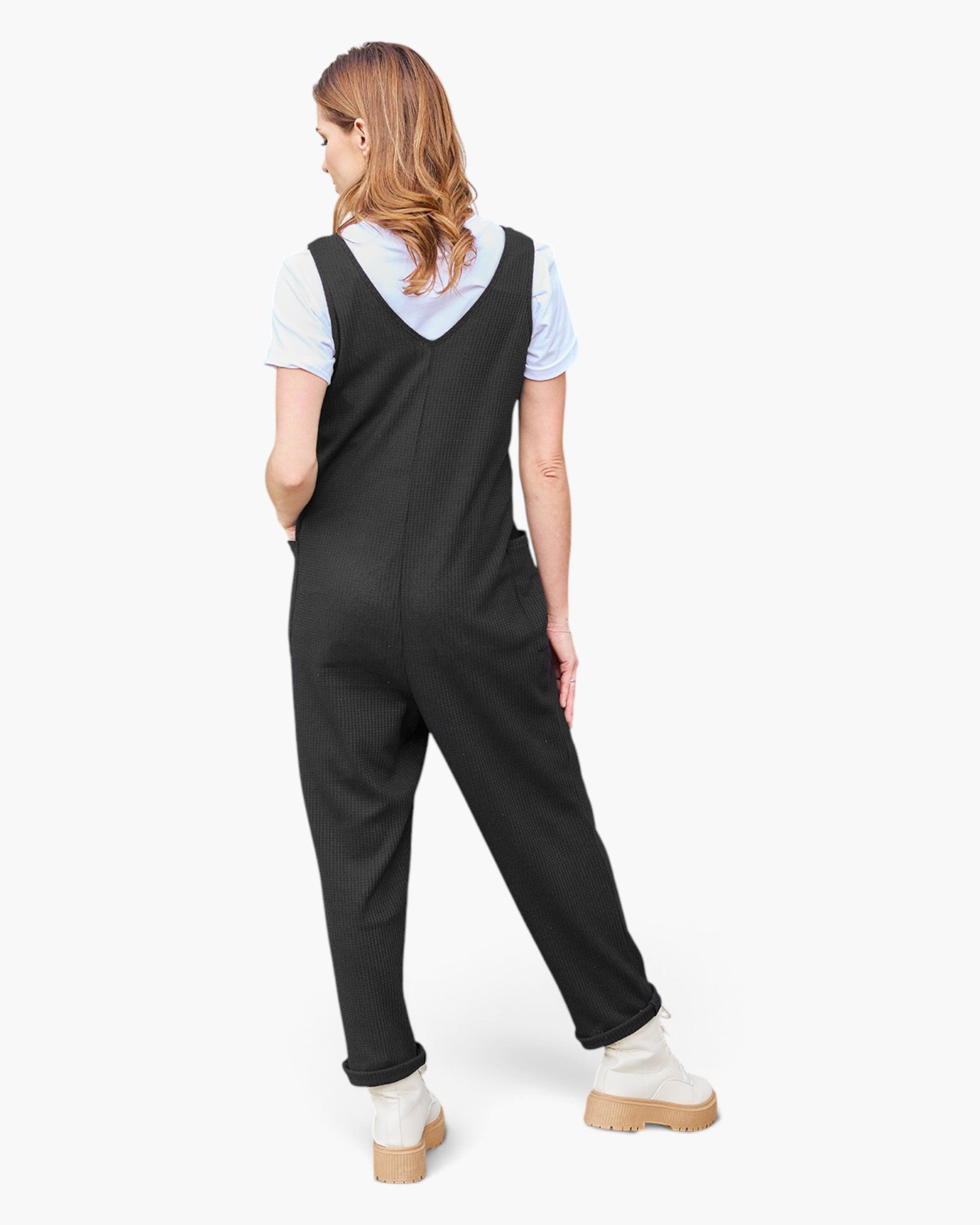 A woman shown from the back wearing a black sleeveless jumpsuit over a light blue short-sleeve shirt, paired with white platform boots, against a plain white studio background.
