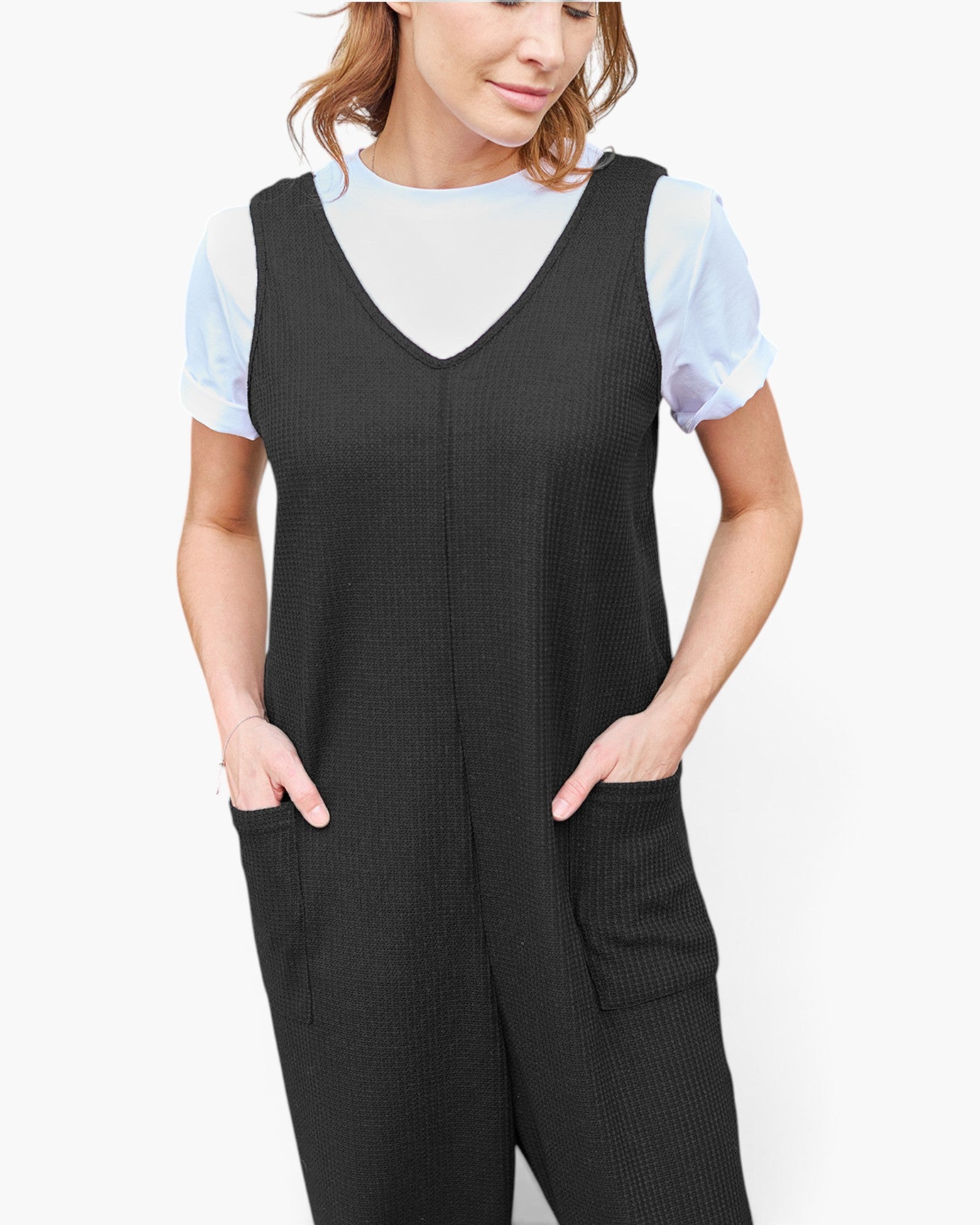A woman wearing a black ribbed sleeveless jumpsuit with front patch pockets over a light blue short-sleeve top, photographed against a plain light gray studio background.