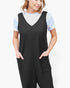 A woman wearing a black ribbed sleeveless jumpsuit with front patch pockets over a light blue short-sleeve top, photographed against a plain light gray studio background.