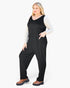 A woman modeling a black sleeveless jumpsuit with side pockets over a light gray long-sleeve shirt and brown platform shoes, photographed against a plain white studio background.