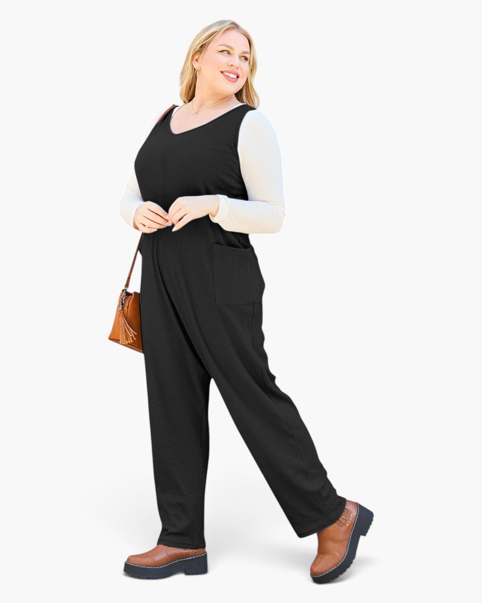 A woman modeling a black sleeveless jumpsuit with wide legs, styled over a white long-sleeve top and brown ankle boots, holding a small brown handbag against a plain white background.