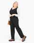 A woman modeling a black sleeveless jumpsuit with wide legs, styled over a white long-sleeve top and brown ankle boots, holding a small brown handbag against a plain white background.