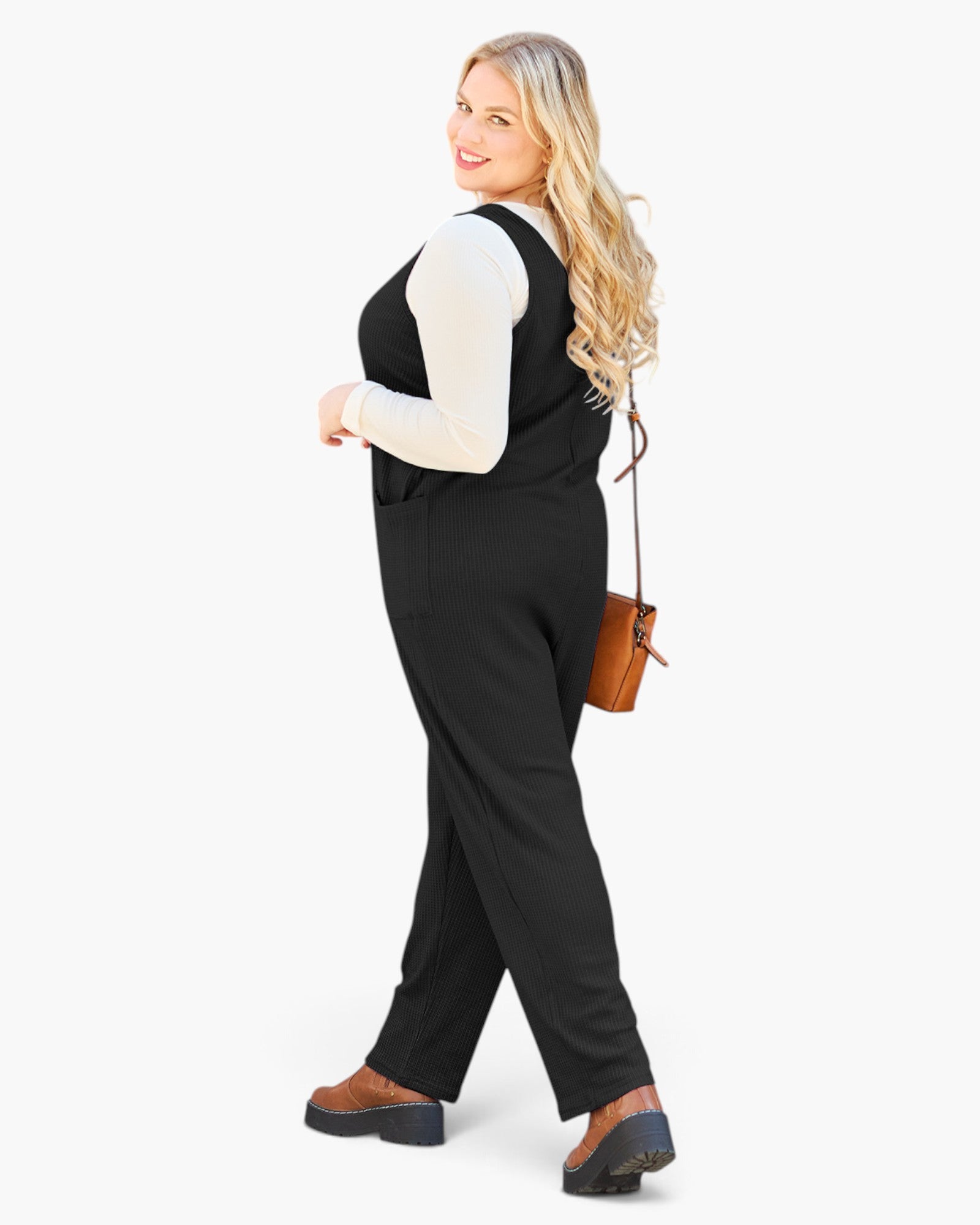 A woman modeling a black sleeveless jumpsuit over a white long-sleeve top, styled with brown platform shoes and a small brown crossbody bag, against a plain white background.
