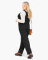 A woman modeling a black sleeveless jumpsuit over a white long-sleeve top, styled with brown platform shoes and a small brown crossbody bag, against a plain white background.