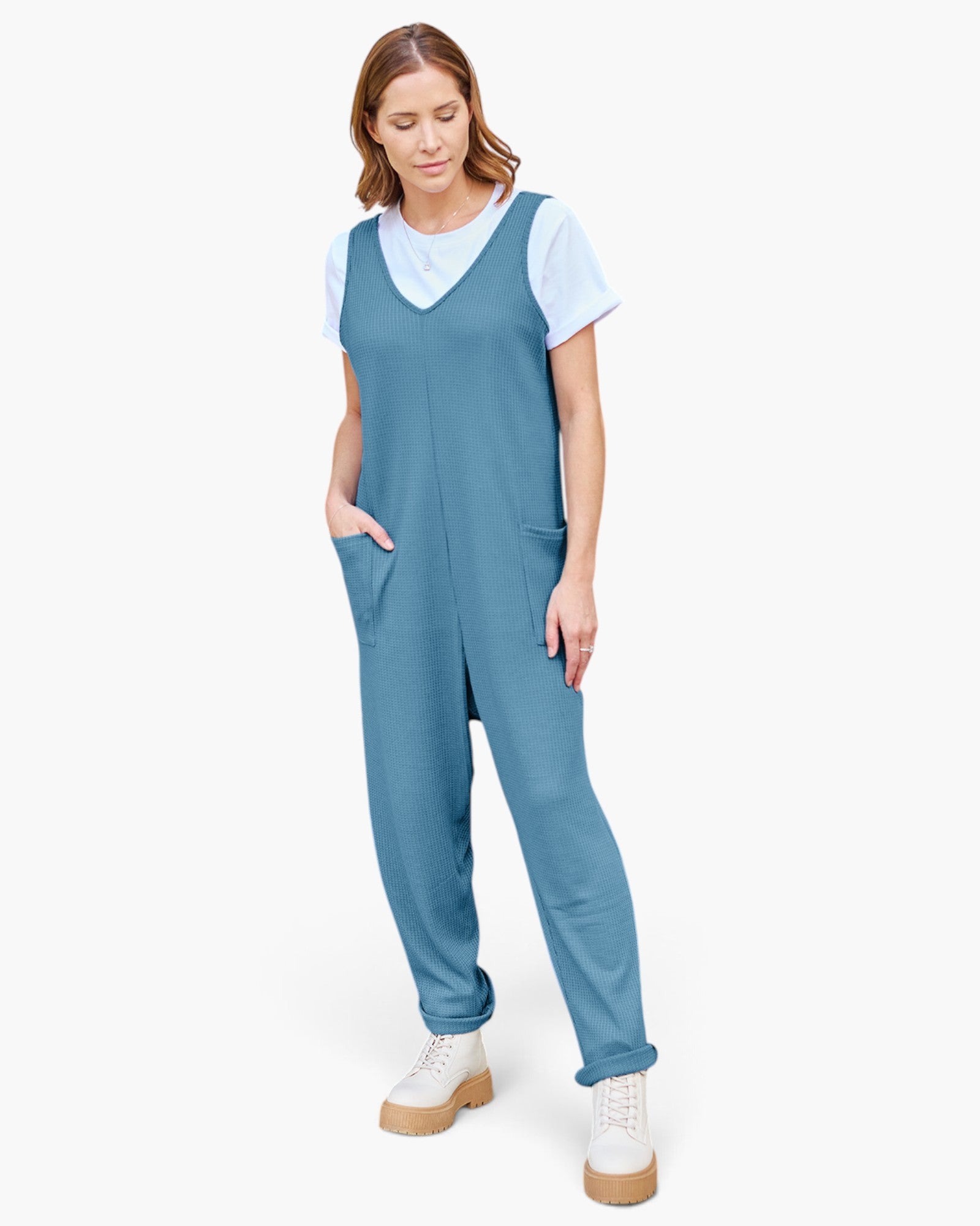 A woman modeling a slate blue sleeveless jumpsuit over a white t-shirt and white platform sneakers, photographed against a solid off-white background.