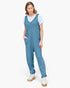 A woman modeling a slate blue sleeveless jumpsuit over a white t-shirt and white platform sneakers, photographed against a solid off-white background.
