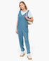 A woman modeling a slate blue sleeveless jumpsuit over a white t-shirt, carrying a beige shoulder bag and wearing white lace-up boots, set against a plain white studio background.