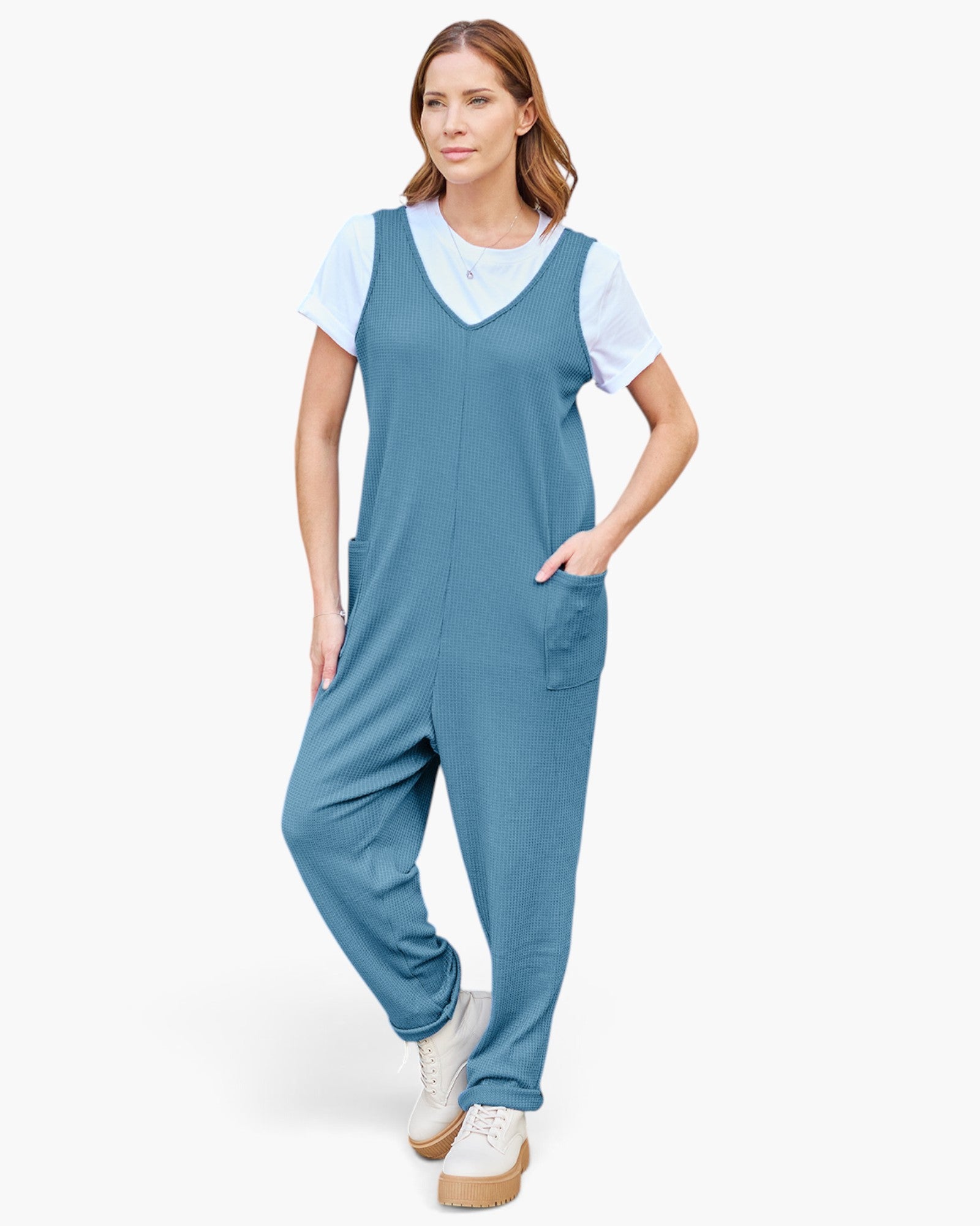 A woman modeling a slate blue sleeveless jumpsuit with a white t-shirt underneath and white platform sneakers, set against a plain white studio background.