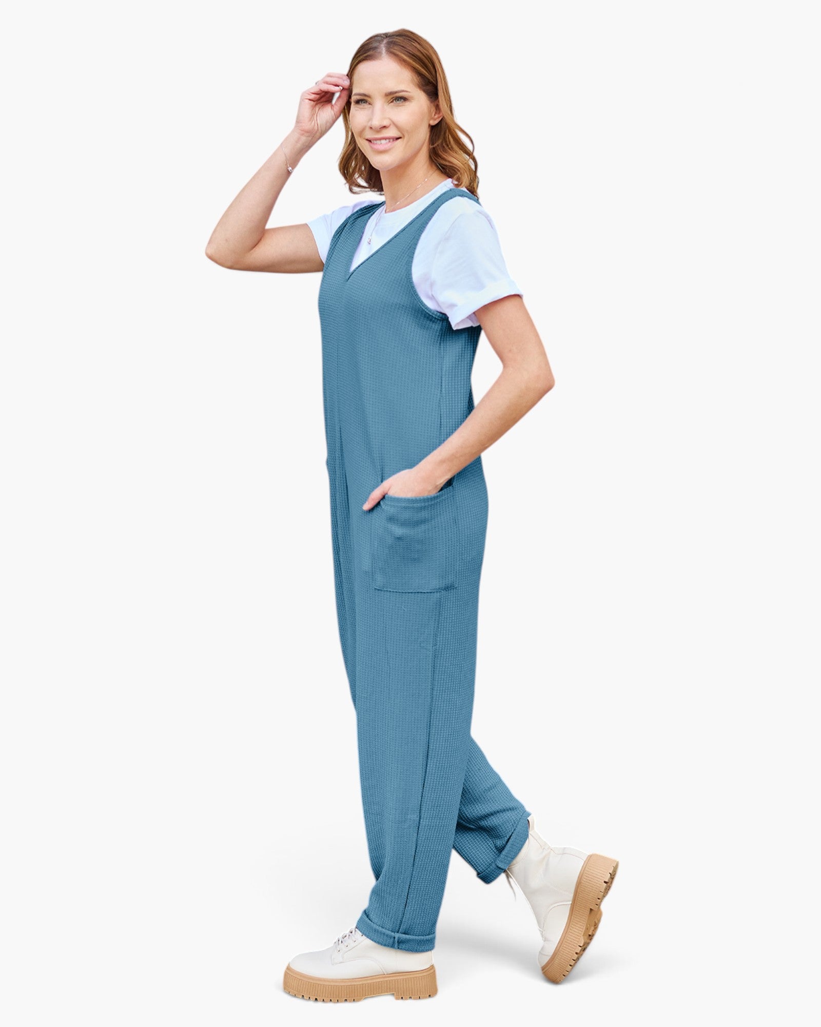 A woman modeling a slate blue scrub jumpsuit over a white short-sleeve V-neck scrub top, paired with white boots, against a solid white background.