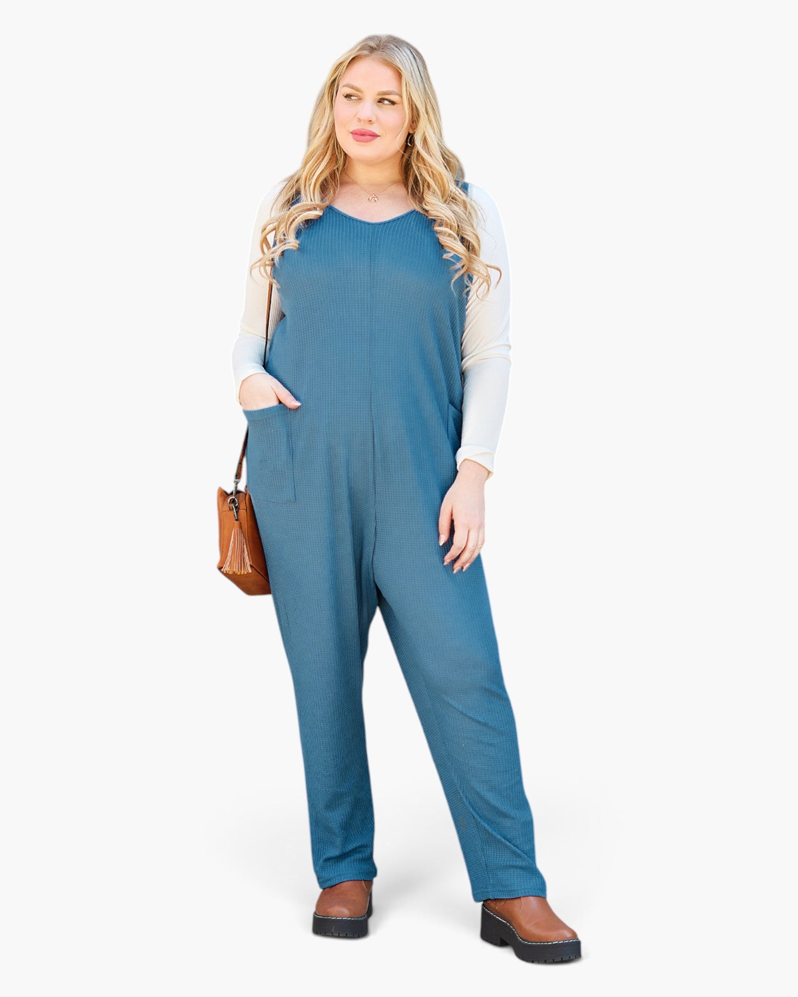 A woman modeling a slate blue sleeveless jumpsuit over a white long-sleeve shirt, holding a brown leather handbag, against a plain white studio background.