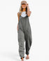 A woman modeling a loose-fitting, sleeveless gray jumpsuit with large front patch pockets, styled with a beige baseball cap, a white tank top, and flat sandals against a plain white background.