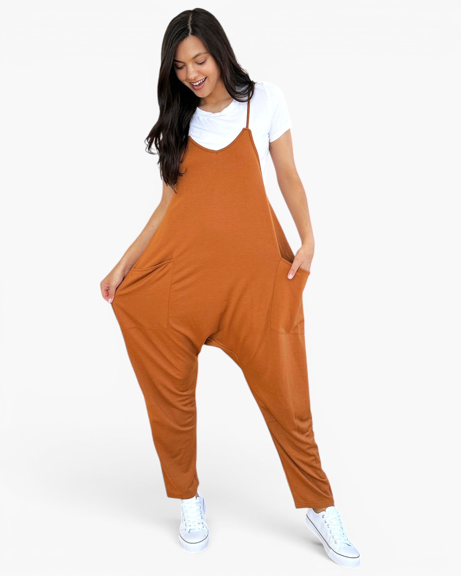 A woman modeling a rust-orange sleeveless jumpsuit with wide legs and front pockets, worn over a white t-shirt and white sneakers, against a light gray studio background.