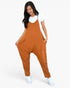 A woman modeling a rust-orange sleeveless jumpsuit with wide legs and front pockets, worn over a white t-shirt and white sneakers, against a light gray studio background.