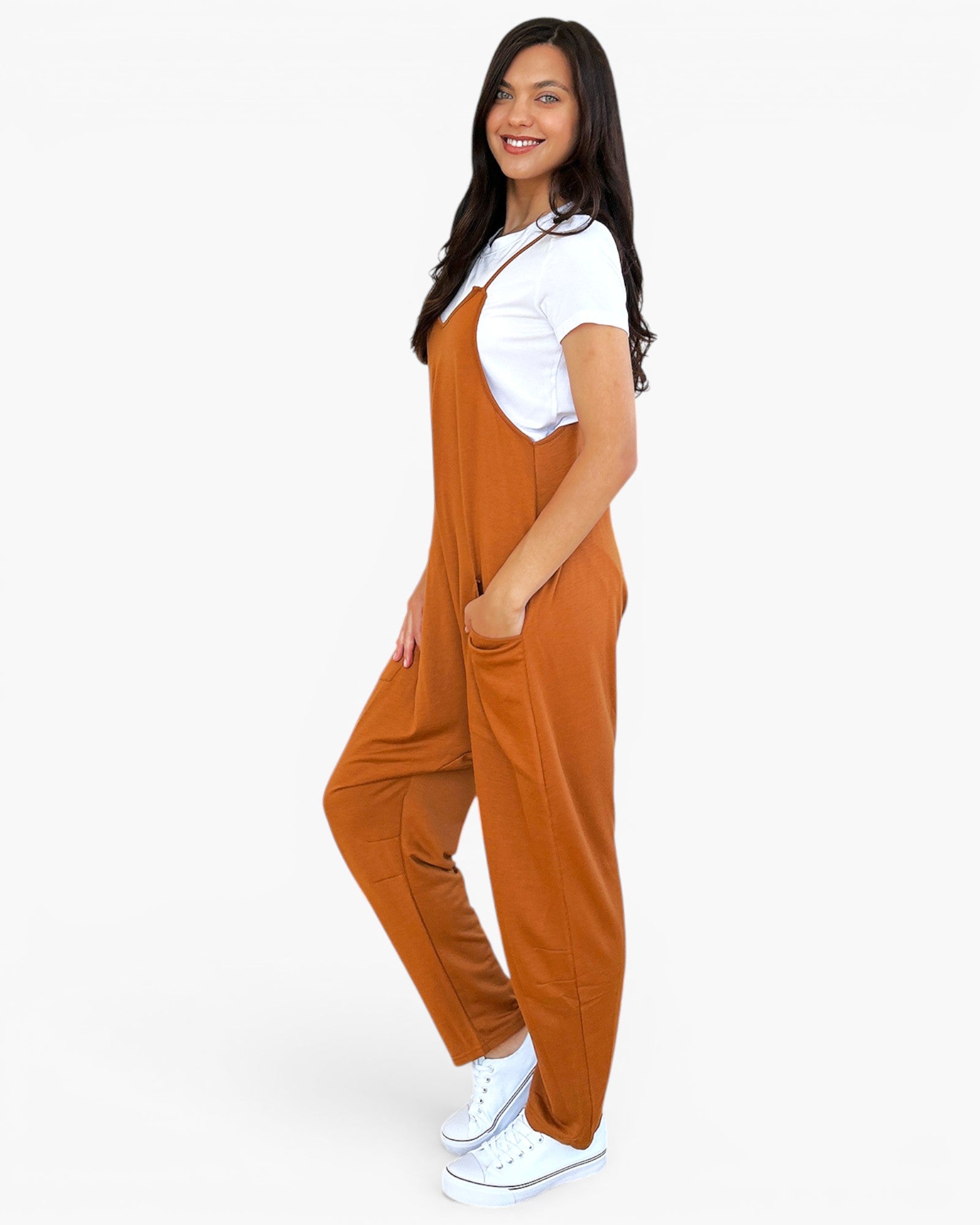 A woman modeling a pair of rust-orange bib jumpsuits over a white t-shirt and white sneakers, standing against a solid white background.
