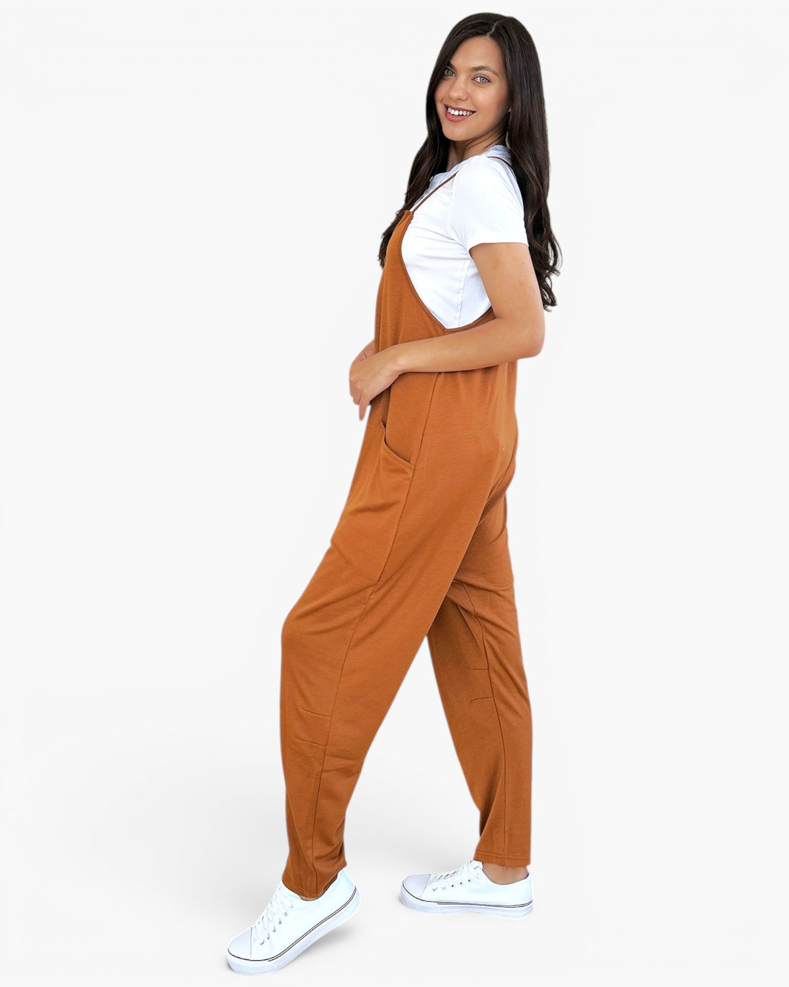 A woman modeling rust-brown overall jumpsuit with side pockets, styled with a white t-shirt and white sneakers, shown from a side-back angle against a plain white studio background.