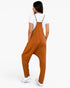 A woman shown from the back wearing a rust-orange harem-style jumpsuit over a white t-shirt and white sneakers, set against a plain white studio background.