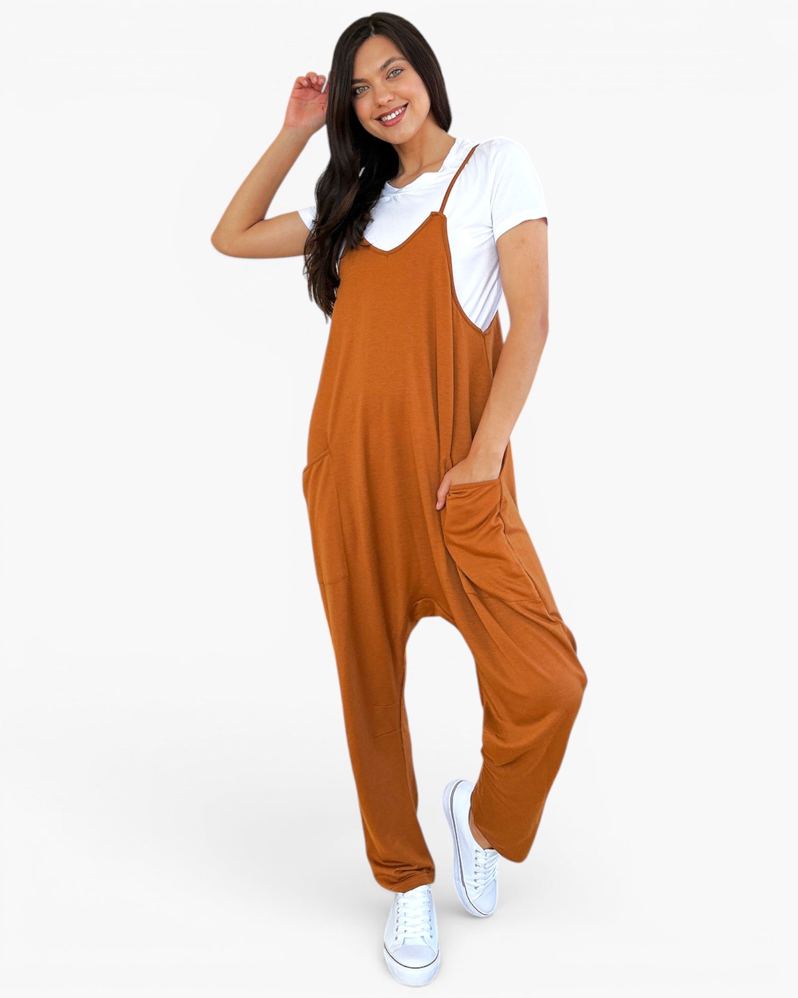 A woman modeling a rust-orange harem-style jumpsuit over a white t-shirt and white sneakers, standing against a plain white studio background.