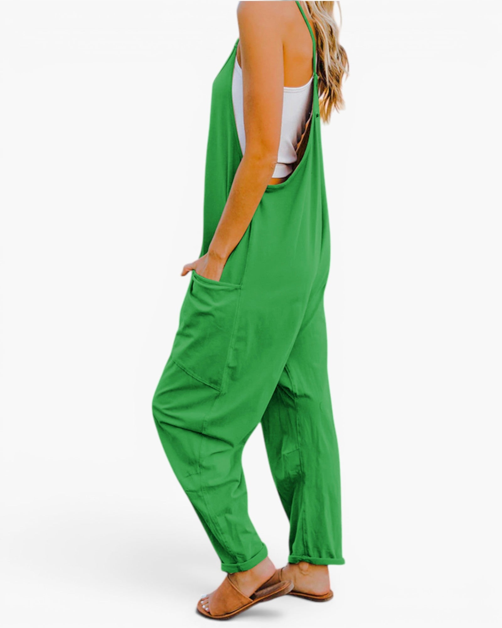 A woman wearing bright green wide-leg jumpsuit overalls with side pockets, styled over a white tank top and brown flat sandals, shown from a side-back angle against a plain white background.