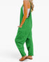 A woman wearing bright green wide-leg jumpsuit overalls with side pockets, styled over a white tank top and brown flat sandals, shown from a side-back angle against a plain white background.