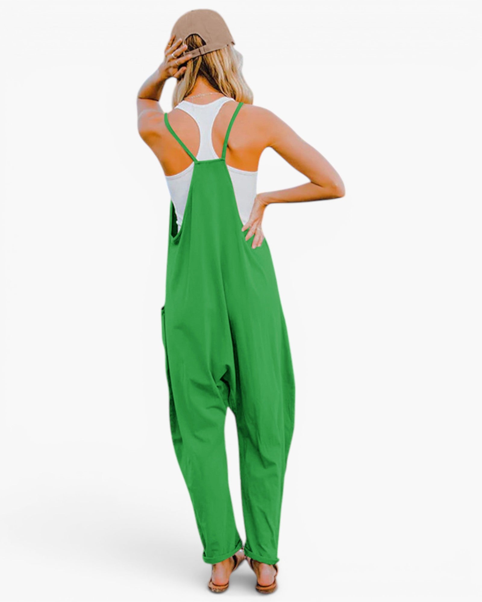 A woman shown from the back wearing bright green overall-style coveralls over a white tank top, with a beige hat and sandals, against a plain light background.