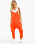 A woman modeling a bright orange sleeveless jumpsuit with a white tank top underneath and white sneakers, standing against a plain white background.