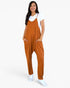 A woman modeling a rust-orange sleeveless jumpsuit with front pockets over a white t-shirt and white sneakers, set against a plain white studio background.