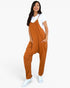 A woman modeling a rust-orange sleeveless jumpsuit with a white t-shirt and white sneakers, standing against a solid white background.
