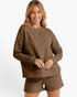 A woman modeling a matching brown textured knit lounge set consisting of a long-sleeve crewneck sweatshirt and matching shorts, against a clean white background.