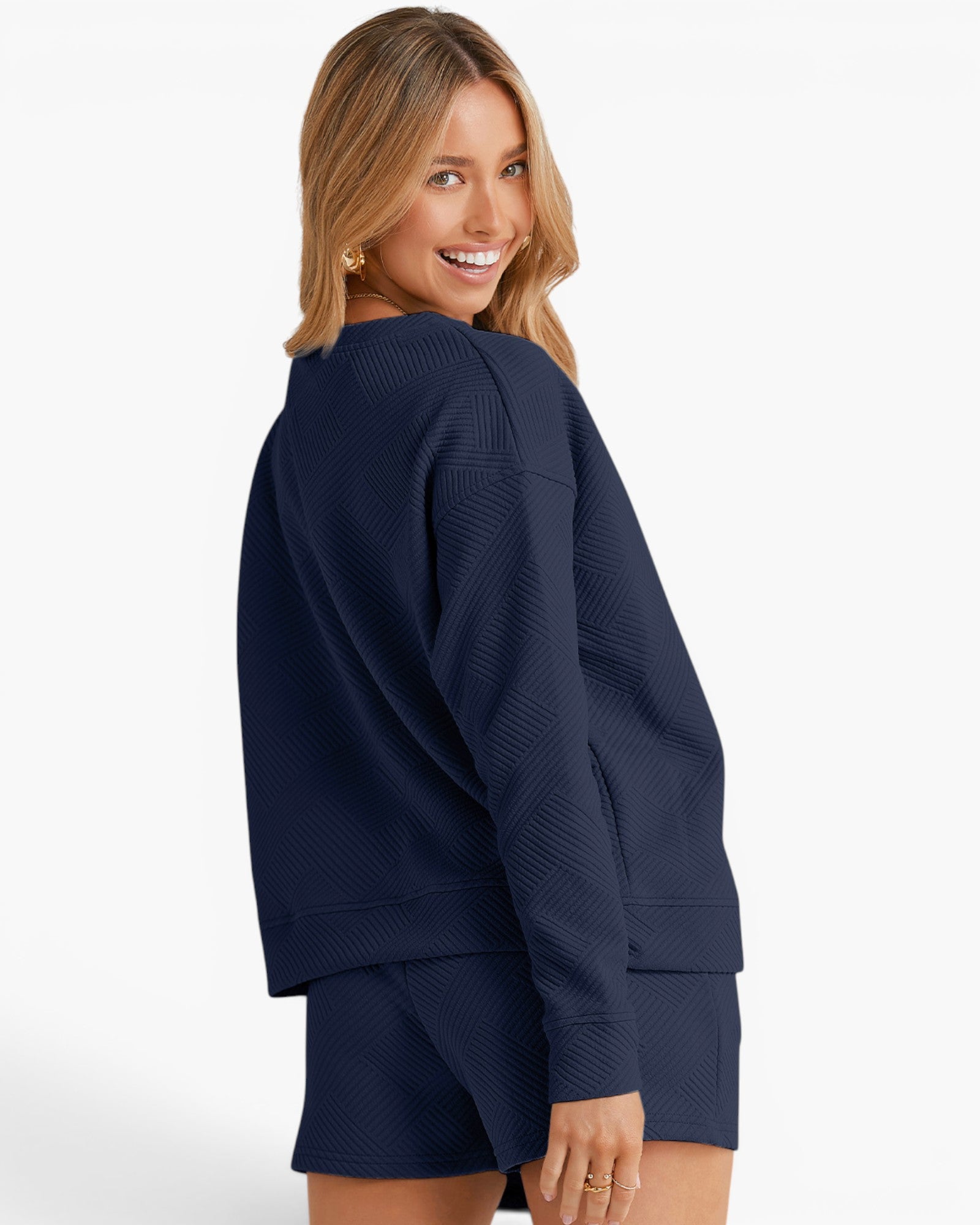 A woman wearing a matching navy blue textured lounge set, featuring a long-sleeve top and shorts, photographed from a three-quarters back angle against a clean white studio background.