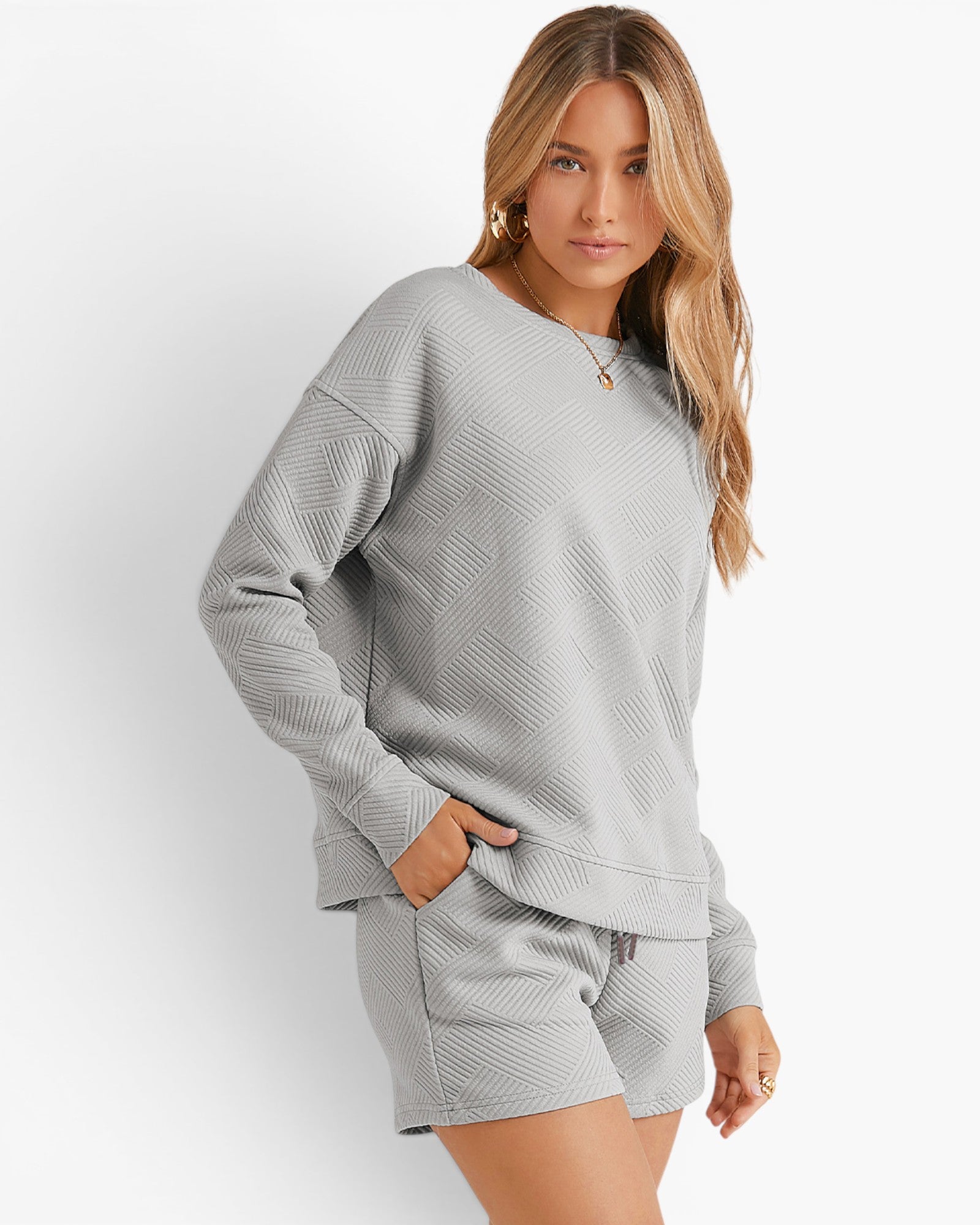 A woman modeling a light gray textured matching lounge set consisting of a long-sleeve sweatshirt and shorts, posed against a clean white studio background.