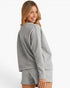 A woman wearing a light gray textured matching lounge set, featuring a long-sleeve sweatshirt and shorts, shown from a three-quarters back view against a plain white studio background.