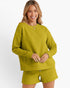 A woman modeling a matching textured knit lounge set in a mustard yellow-green color, featuring a long-sleeve crewneck sweatshirt and coordinating shorts, against a plain white studio background.