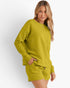 A woman modeling a matching textured knit lounge set in a bright chartreuse yellow-green color, featuring a long-sleeve sweatshirt and shorts, against a plain light gray background.