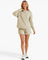 A woman modeling a matching beige lounge set consisting of a long-sleeve sweatshirt and shorts, paired with white sneakers, against a clean white studio background.