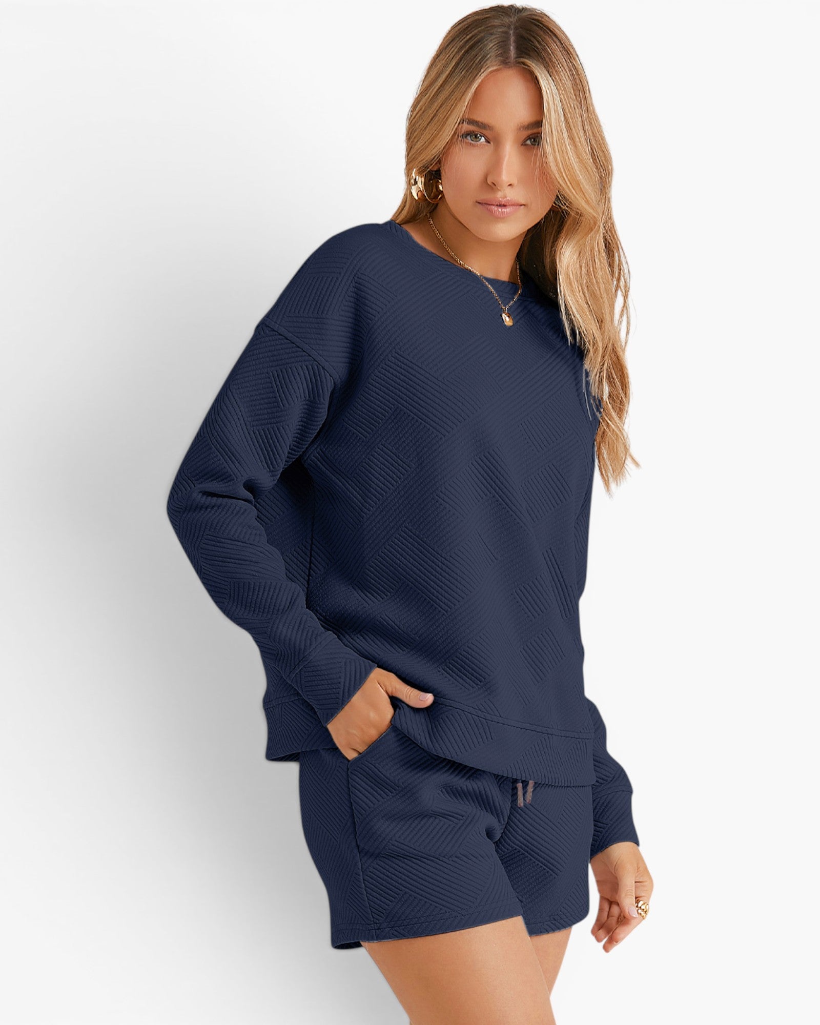 A woman modeling a matching navy blue textured lounge set consisting of a long-sleeve crewneck sweatshirt and shorts, against a clean white studio background.