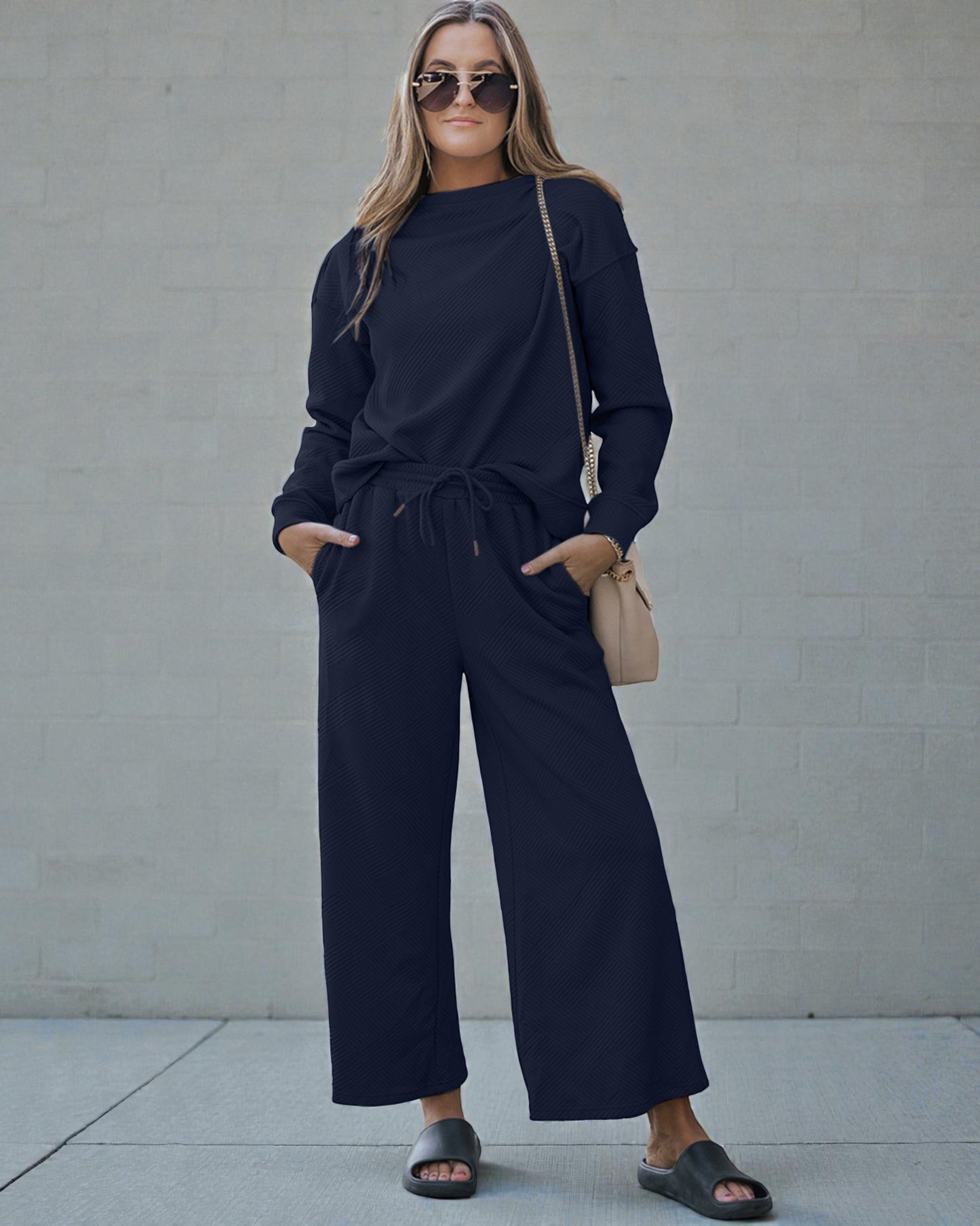Woman wearing a navy blue outfit with sunglasses and a beige handbag against a gray wall.