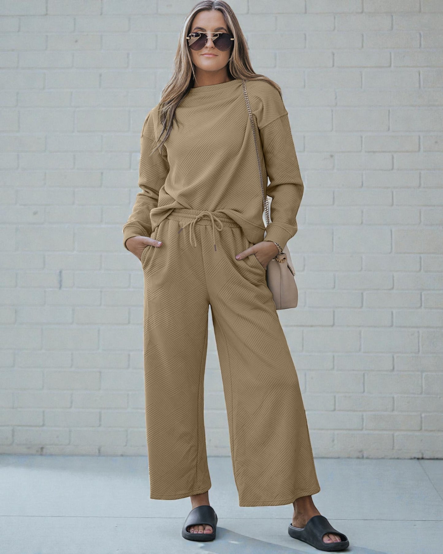 Woman wearing a beige outfit with sunglasses and a handbag against a white brick wall.