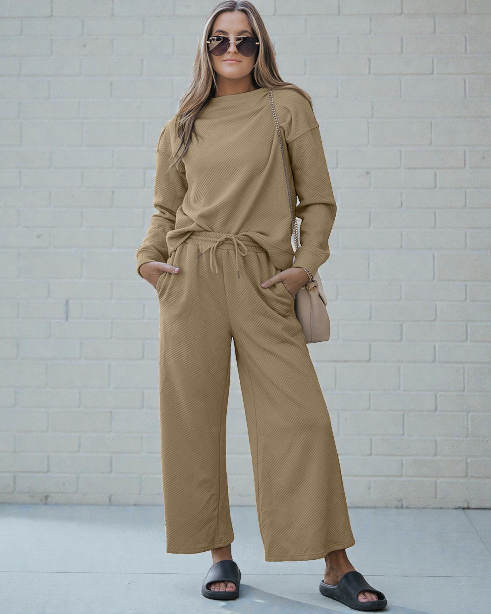 Woman wearing a beige outfit with sunglasses and a handbag against a white brick wall.
