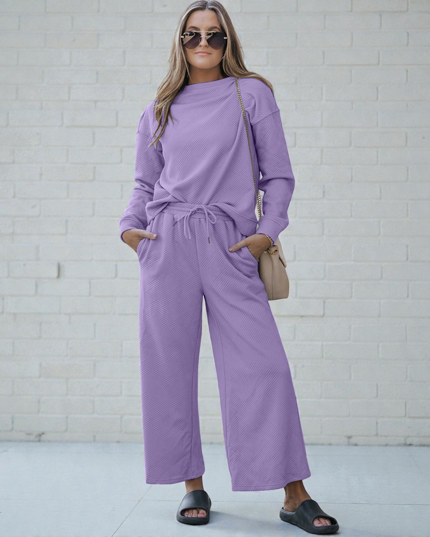 Woman wearing a purple outfit standing against a white brick wall.