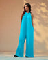 Woman wearing a turquoise jumpsuit standing against a beige wall.