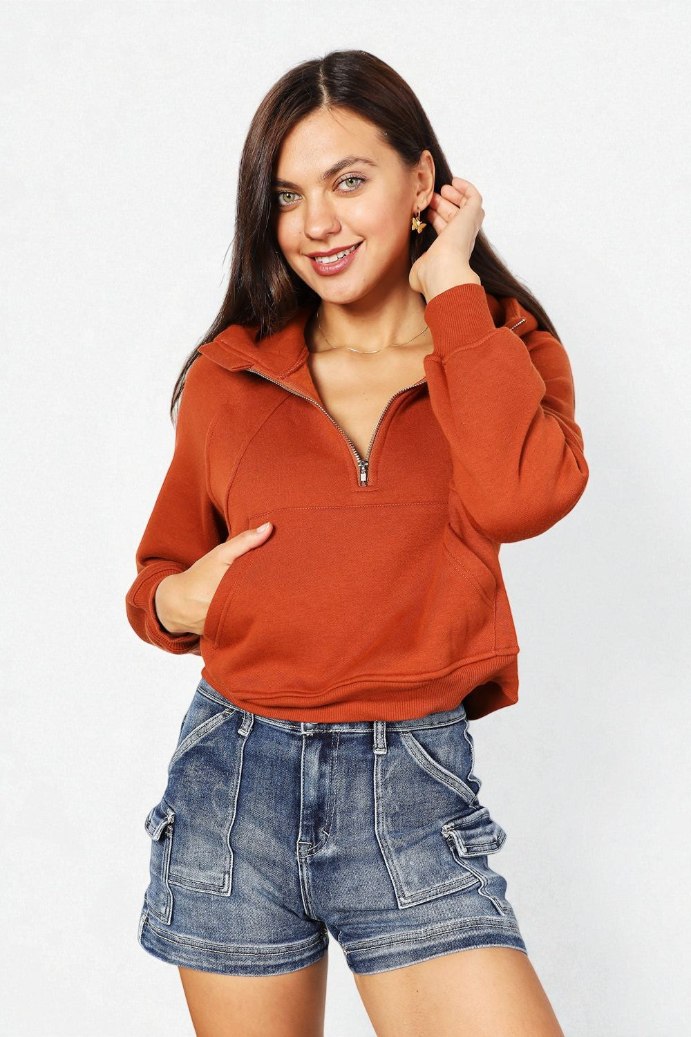 Woman wearing a rust-colored zip-up sweatshirt and denim shorts on a white background