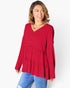 A woman wearing a red tiered long-sleeve blouse with a buttoned V-neckline, paired with blue jeans, photographed against a light gray studio background.