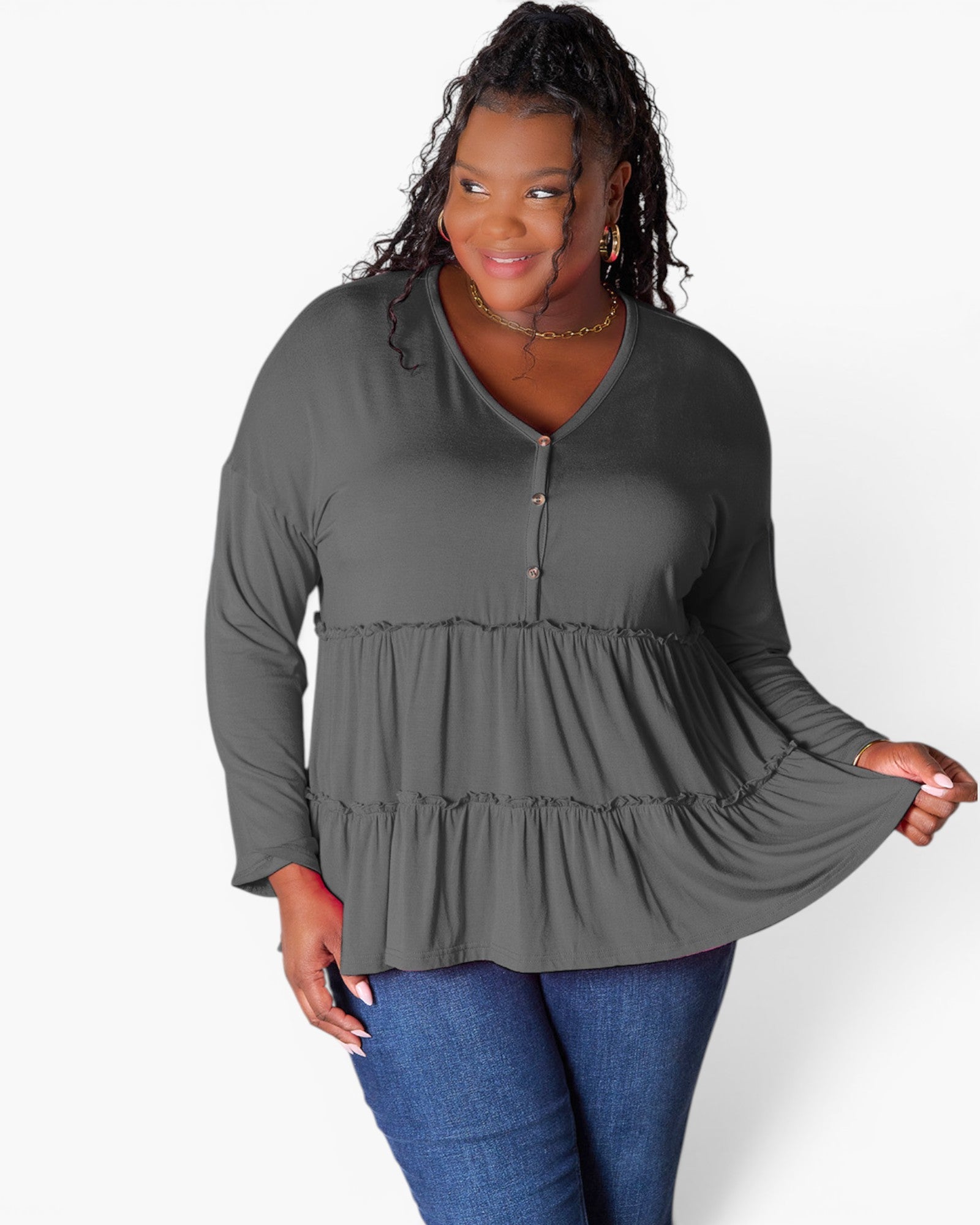 A woman modeling a dark gray long-sleeve top with a tiered peplum waist and a buttoned V-neckline, paired with blue jeans against a plain white background.