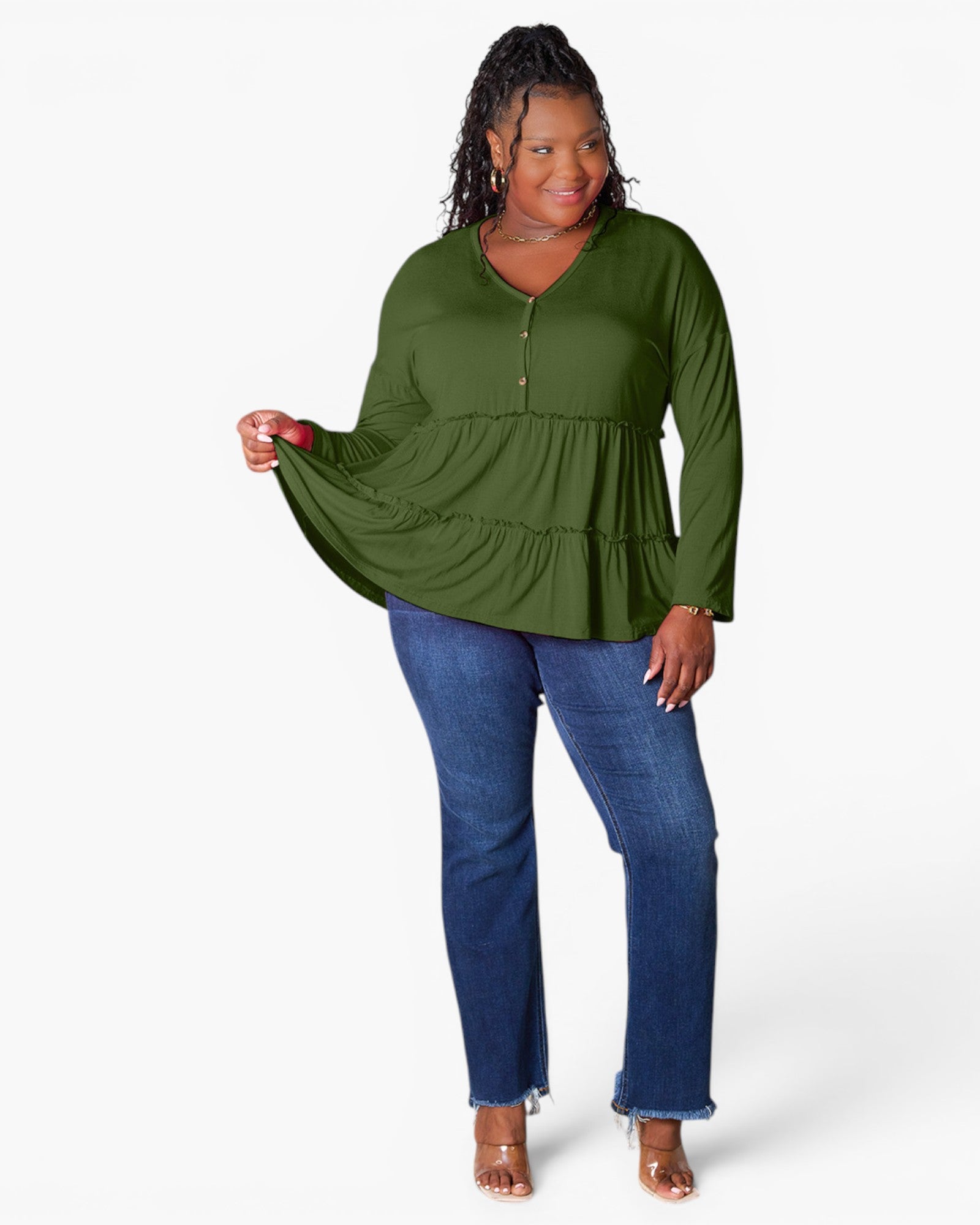 A woman modeling a green long-sleeve tiered peplum top with a half-zip neckline, paired with blue jeans and clear high-heeled sandals, against a plain white studio background.